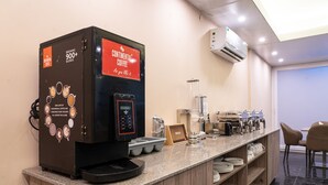 Una estación de café con una máquina de café, una exhibición de pan y un área de asientos con sillas.