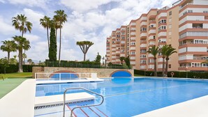 A swimming pool with a diving board, surrounded by palm trees and a multi-story building.