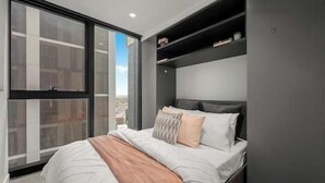 A modern bedroom with a large window, a bed with a beige blanket, and a built-in shelf above the bed.