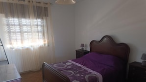 Ein Schlafzimmer mit einem hölzernen Bett, einem Nachttisch mit Lampe, einer Kommode und einem Fenster mit Vorhängen.