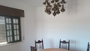 Ein Essbereich mit einem runden Tisch, vier Stühlen, einer Kandelaber, einem Fenster mit Jalousien und einem Fliesenboden.