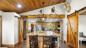 Une cuisine spacieuse dotée d’un îlot central, de tabourets de bar en bois et d’un plafond avec des poutres apparentes et un luminaire suspendu.