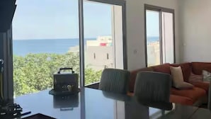 Une pièce moderne avec une grande table et deux chaises, de grandes fenêtres donnant sur la mer et un canapé rouge.