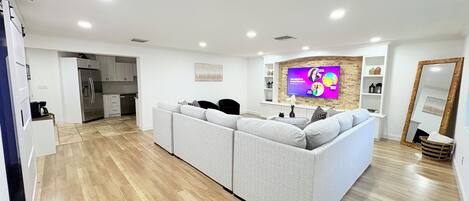 A modern living room with a grey sofa, a flat-screen TV, and a kitchen in the background.