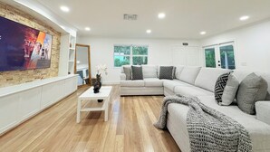 Een moderne woonkamer met een grote flatscreen televisie aan een bakstenen muur, een witte bank met decoratieve kussens, een witte salontafel en een groot raam dat voor natuurlijk licht zorgt.