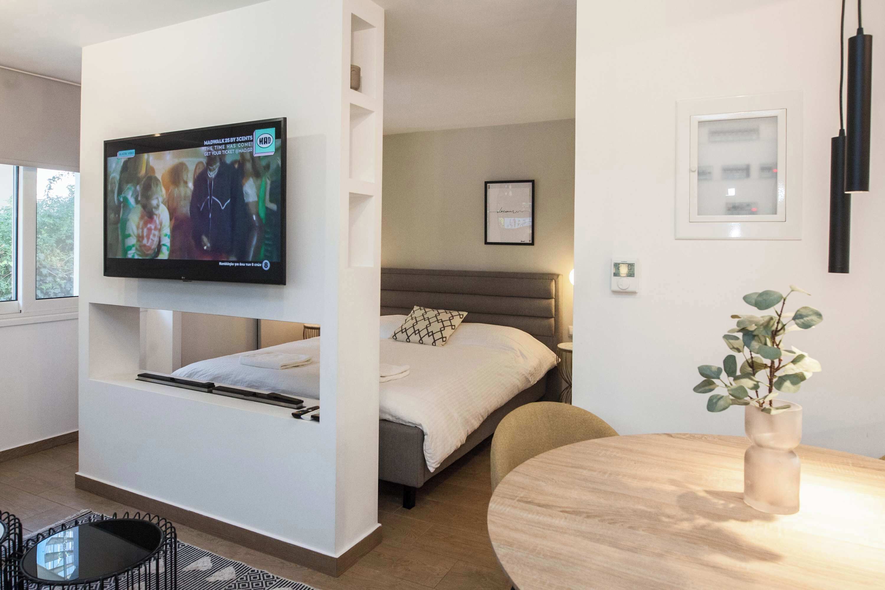 A modern hotel room with a bed, a TV mounted on a wall, a small table, and a plant.