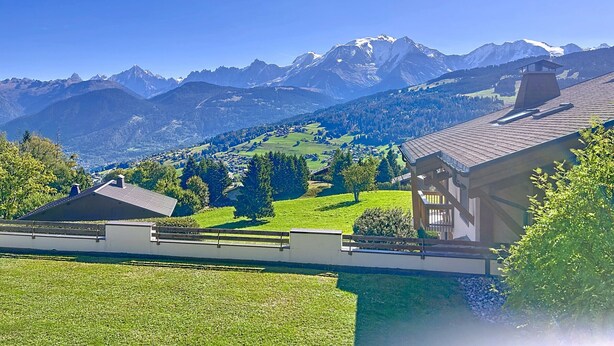 A mountain landscape with a house featuring a large balcony and a view of the surrounding hills and mountains.