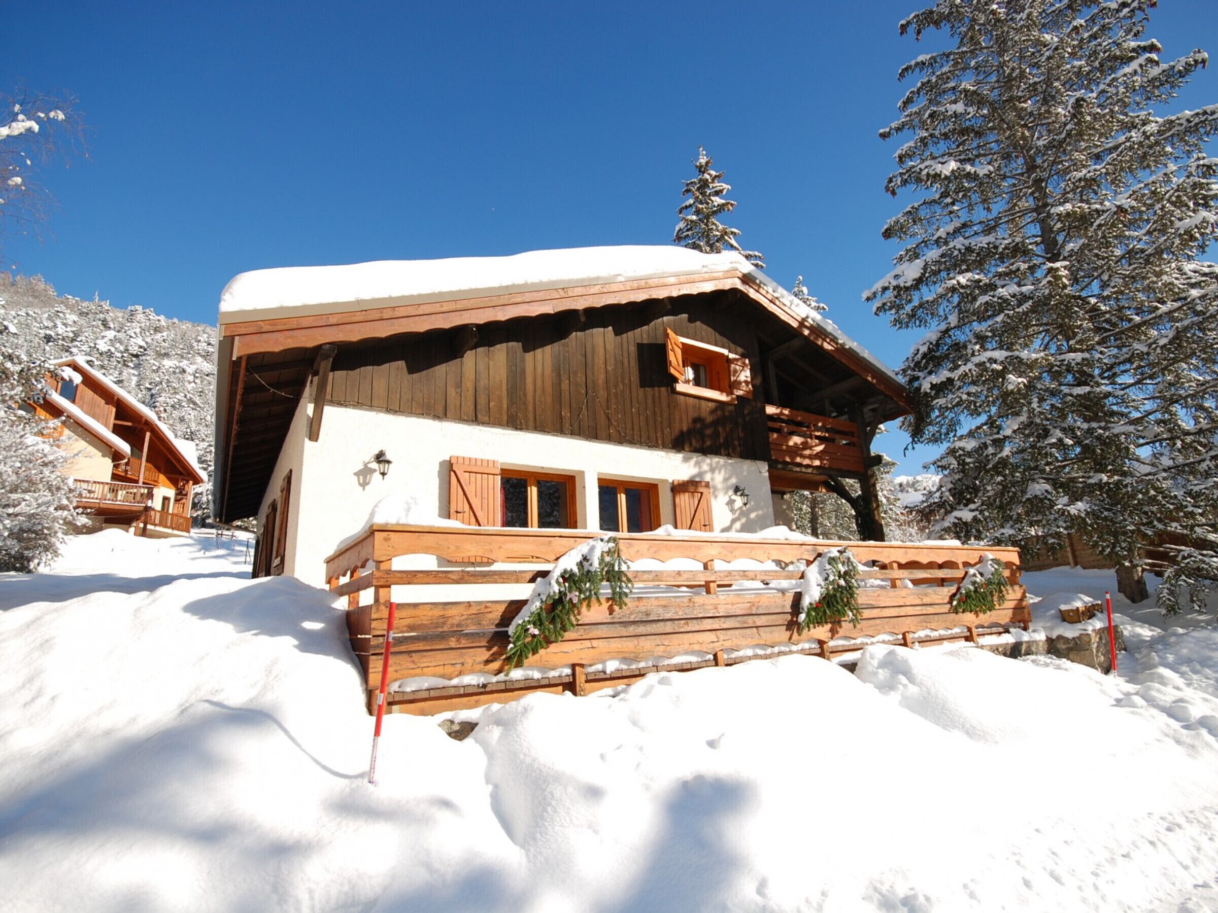 A wooden chalet with a sloped roof and wooden balcony, surrounded by snow-covered trees and a clear blue sky.
