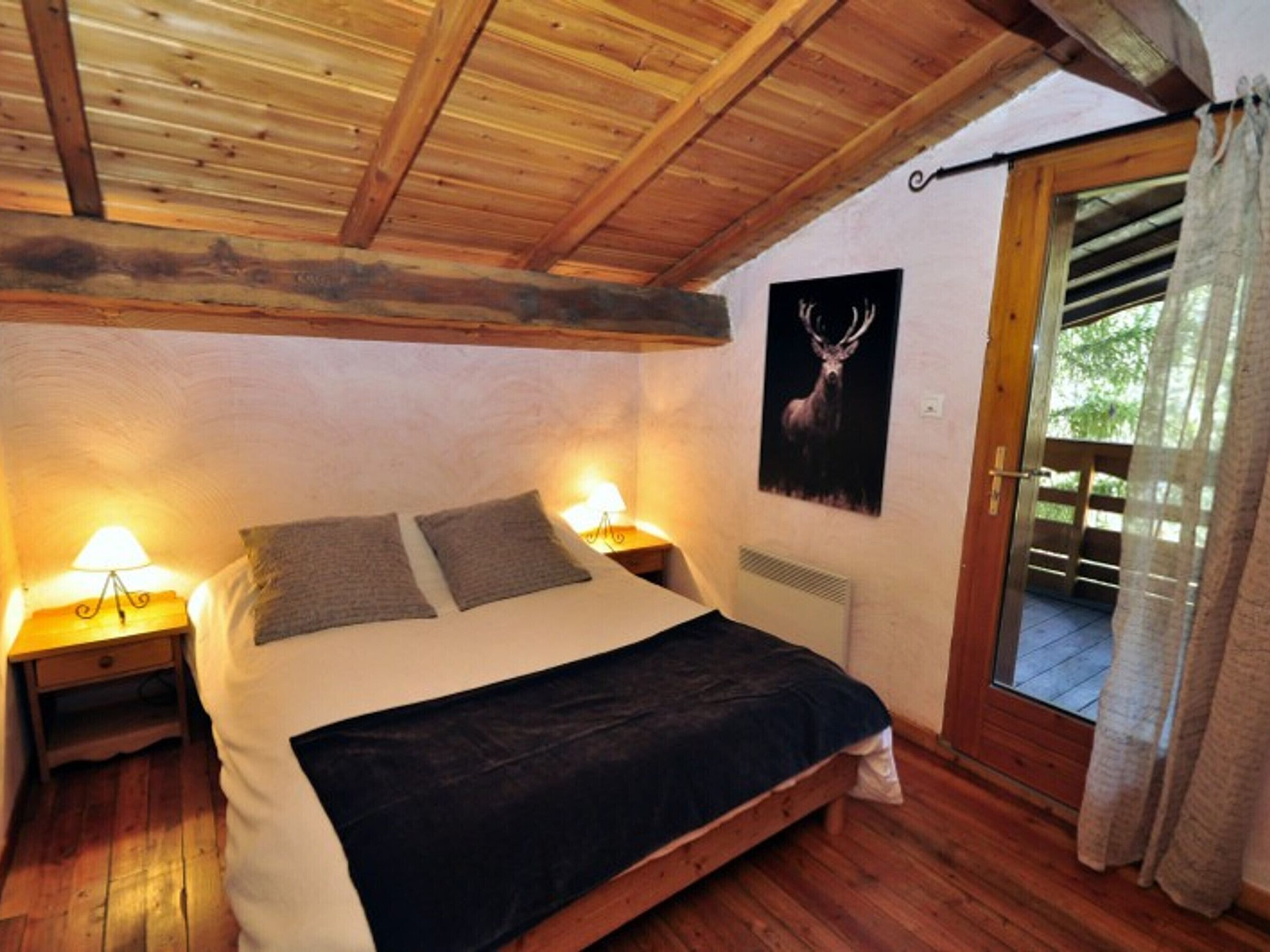 A bedroom with a wooden ceiling, a bed with a dark bedspread, two bedside lamps, a poster of a deer, and a sliding glass door leading to an outdoor area.