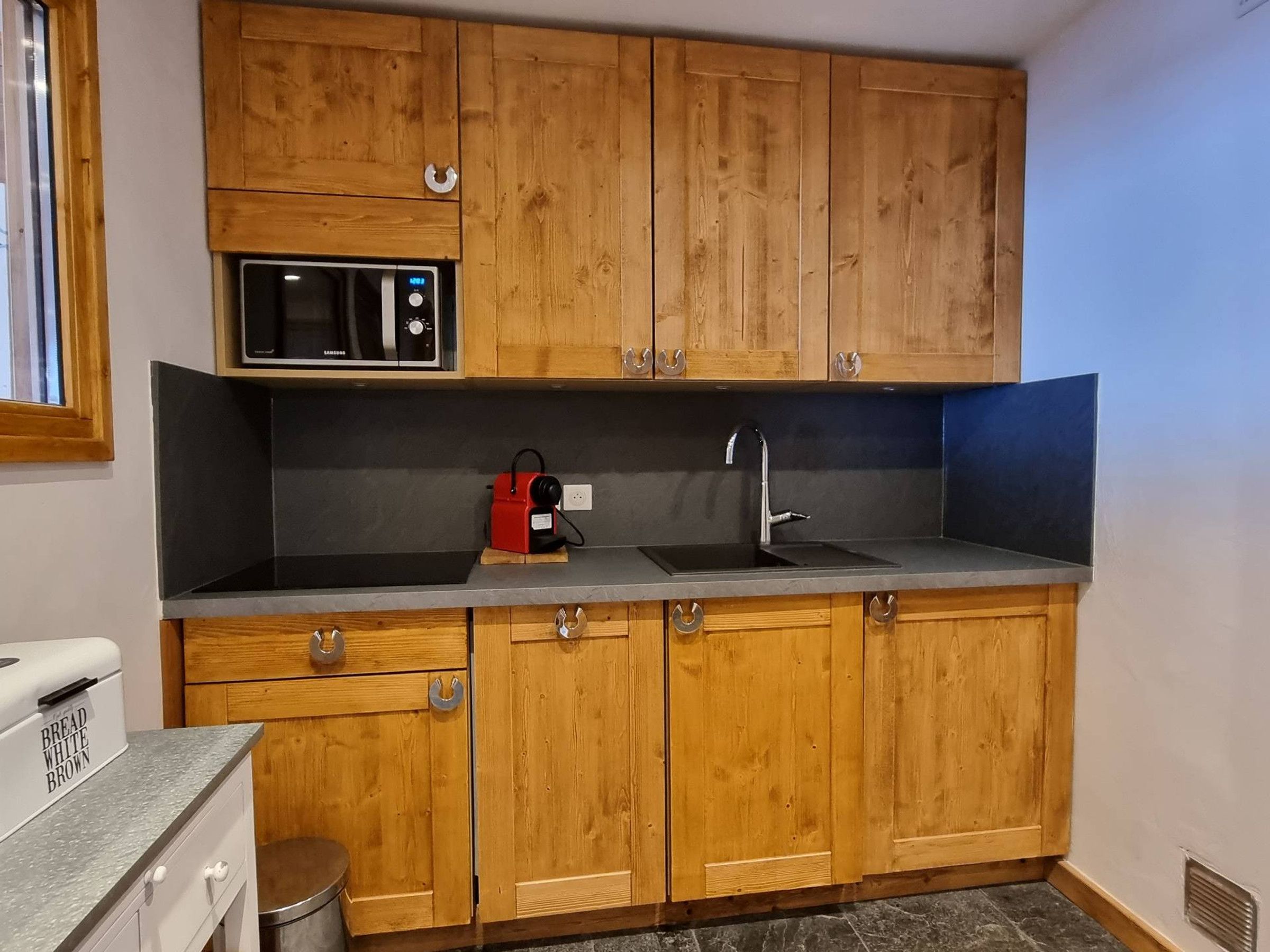 Een keuken met houten kastjes, een magnetron, een spoelbak en een broodmachine.