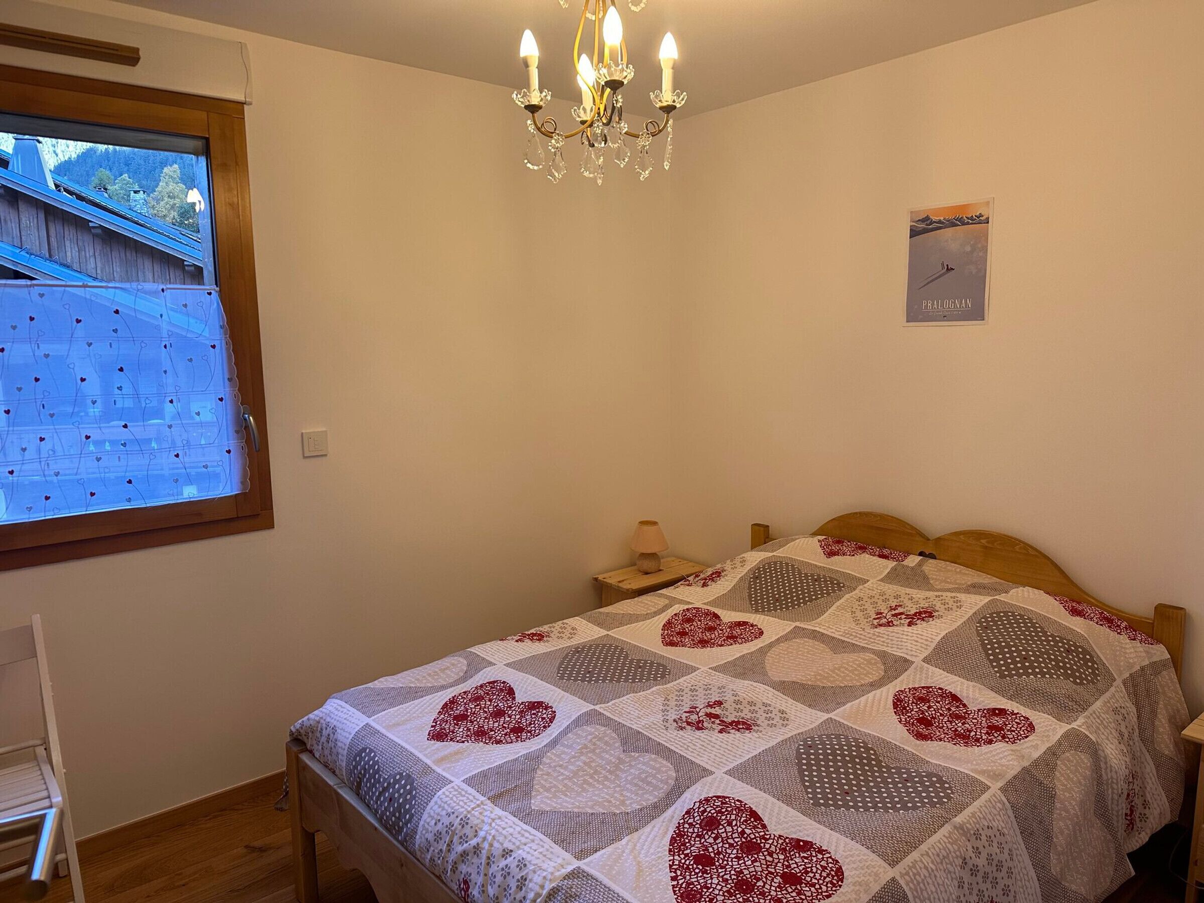 Une chambre avec un lit en bois, une courtepointe ornée de motifs en forme de cœur, un lustre, une fenêtre donnant sur une maison et un panneau accroché au mur.