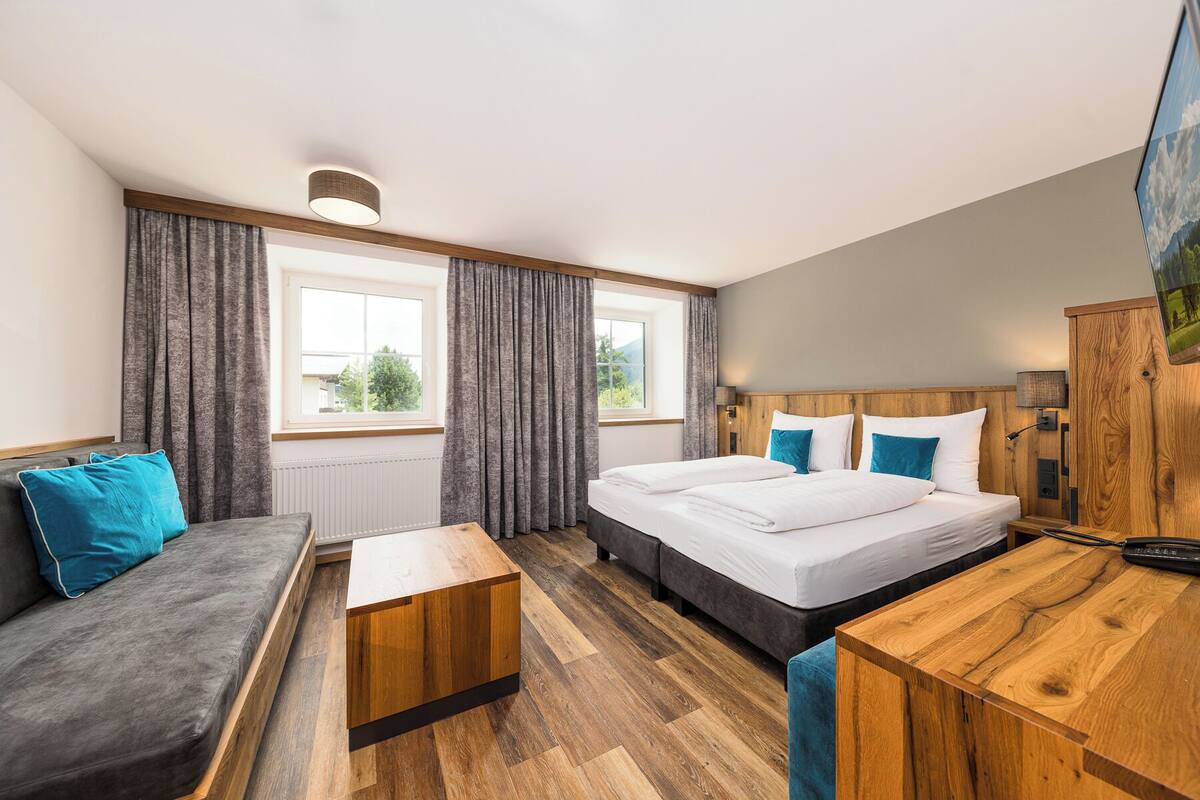 A modern hotel room with a sofa, a wooden desk, a bed with white bedding, and a view of greenery through the windows.