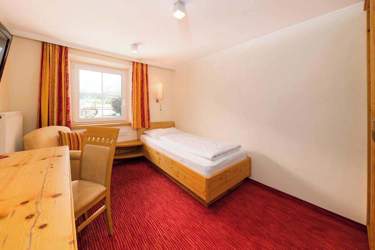A hotel room with a single bed, a wooden desk, a chair, and a window with curtains.