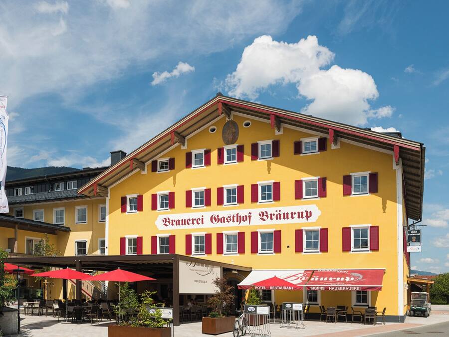 Un edificio amarillo con contraventanas rojas y un letrero que dice 'Bräuerei Gasthof Bräuerei'. Hay mesas y sombrillas al aire libre, y una bandera con el texto 'Bräuerei Gasthof Bräuerei' es visible.