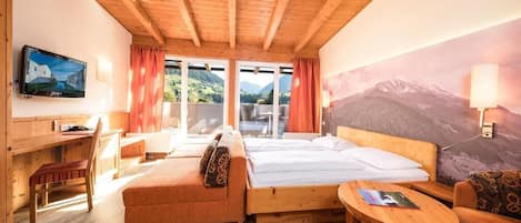 A wooden cabin room with a bed, sofa, desk, and a view of mountains through large windows.