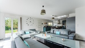 A modern living room with a glass coffee table, grey sofas, and a kitchen in the background. - Holiday Home in the Heart of the Basin with Pool (Cazaux)