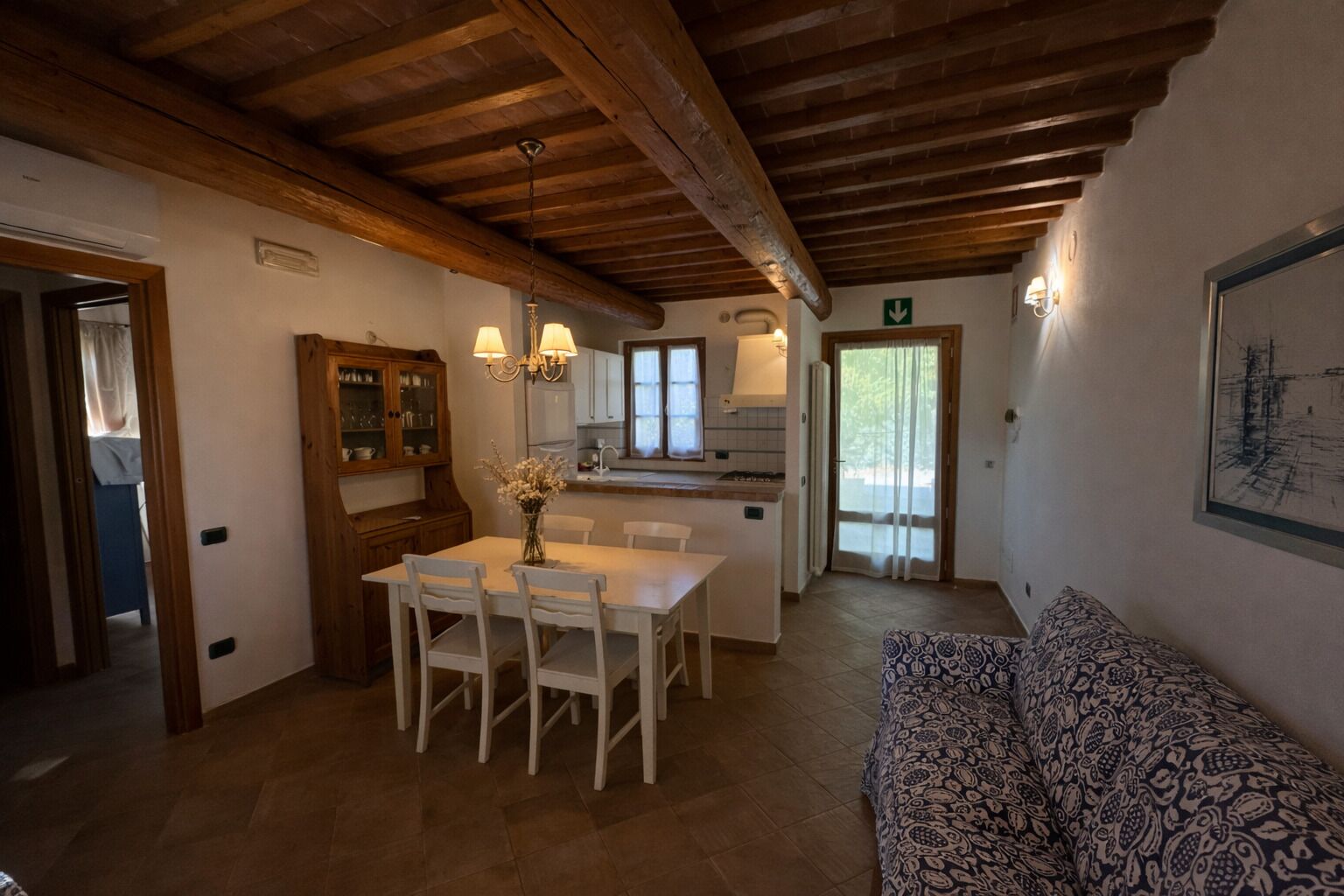 A dining area with a wooden table and chairs, a kitchen with cabinets and a window, and a patterned sofa.