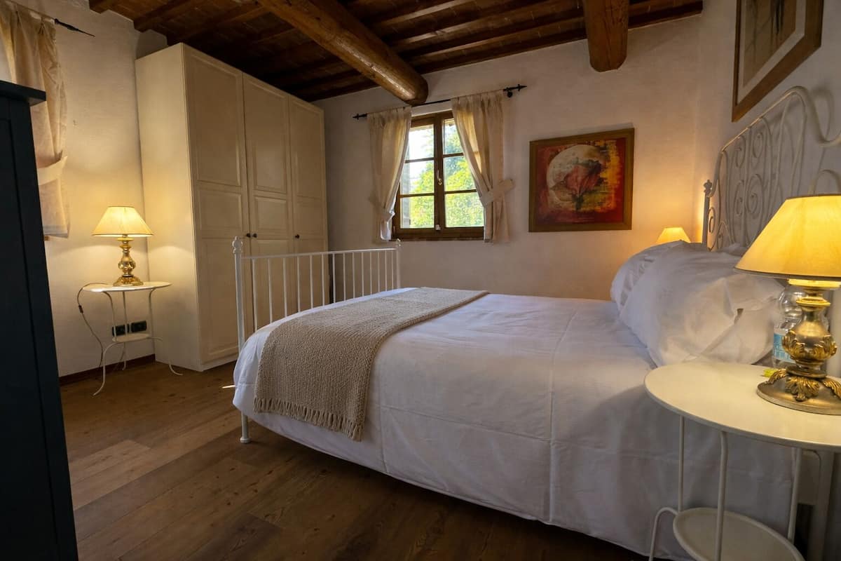 A bedroom with a bed, bedside table, lamp, window, and a painting on the wall.