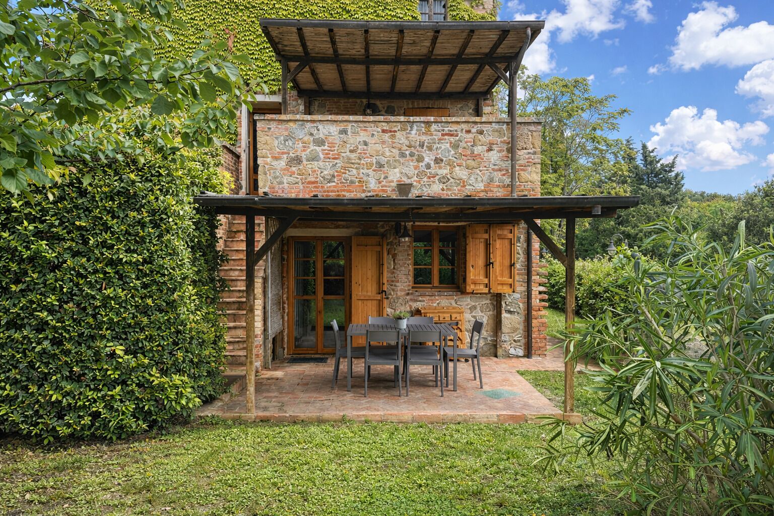 A stone building with a wooden pergola, a table with chairs, and a garden.