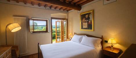 A bedroom with a bed, a nightstand, a lamp, a window with a view, and a radiator.