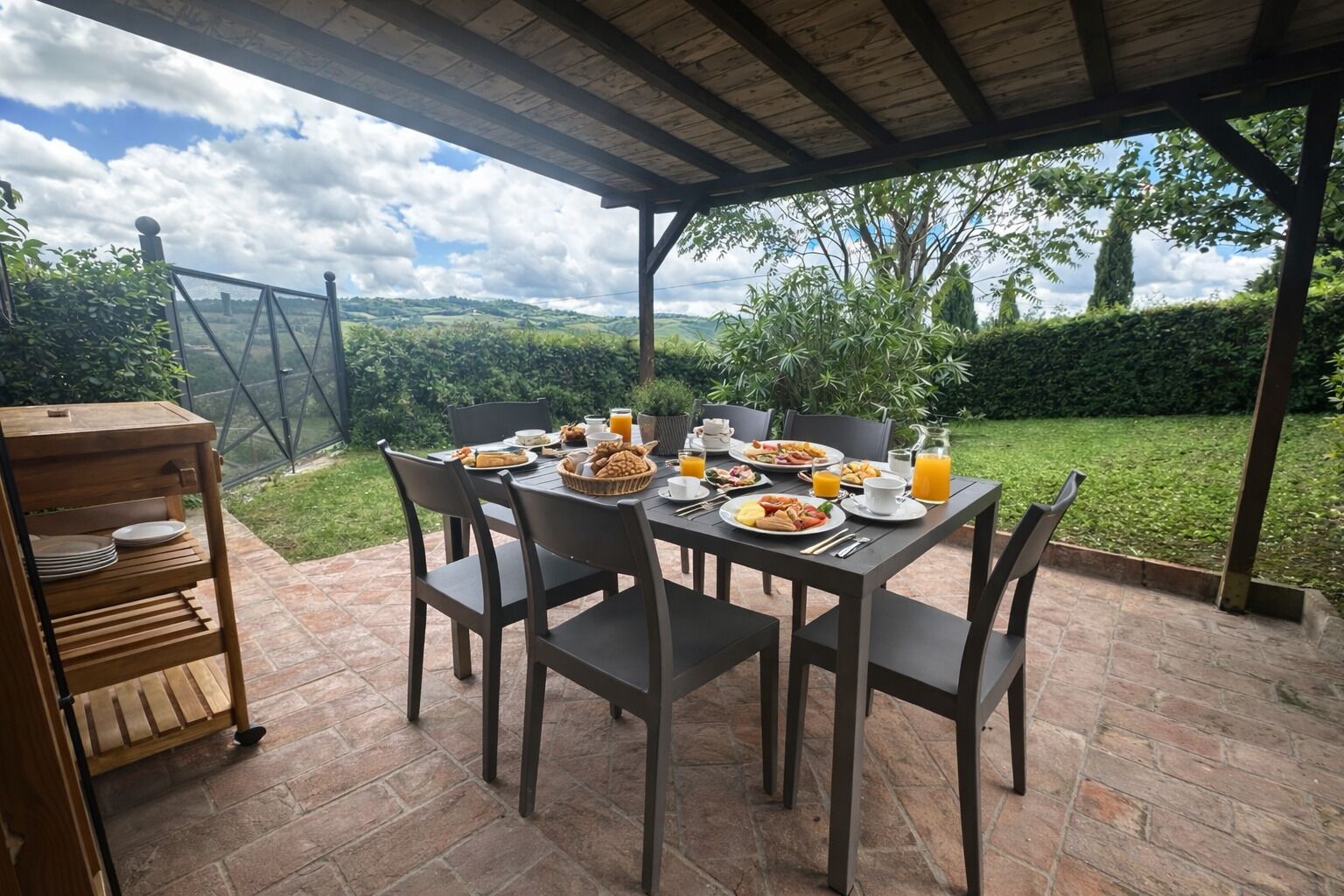 An outdoor patio with a table set for breakfast, surrounded by chairs, and a wooden sideboard.