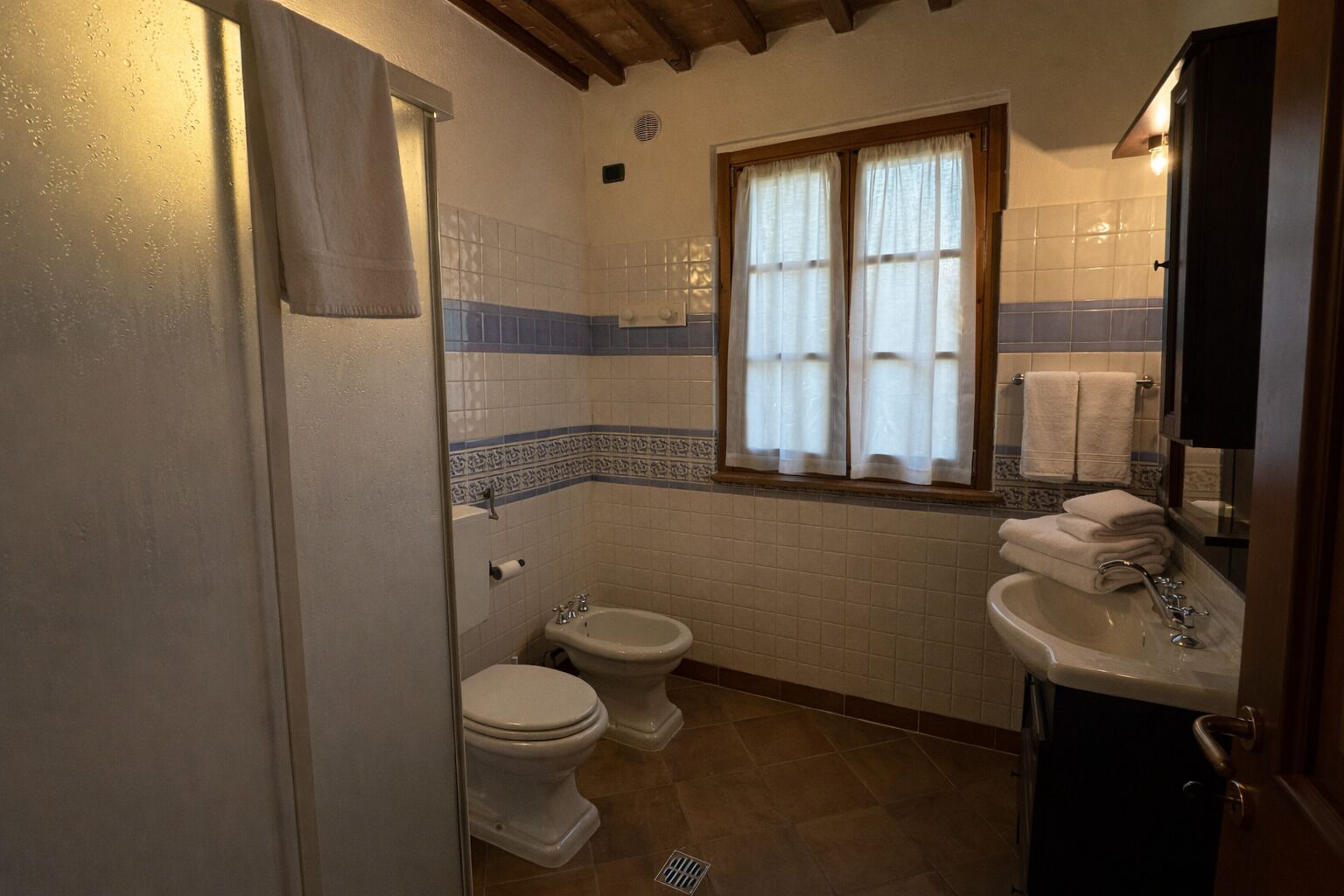 A bathroom with two toilets, a bidet, a sink, and a window with curtains.