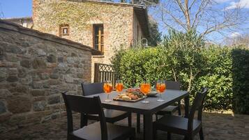 An outdoor dining area with stone walls, a table set with chairs, and a view of a stone building.