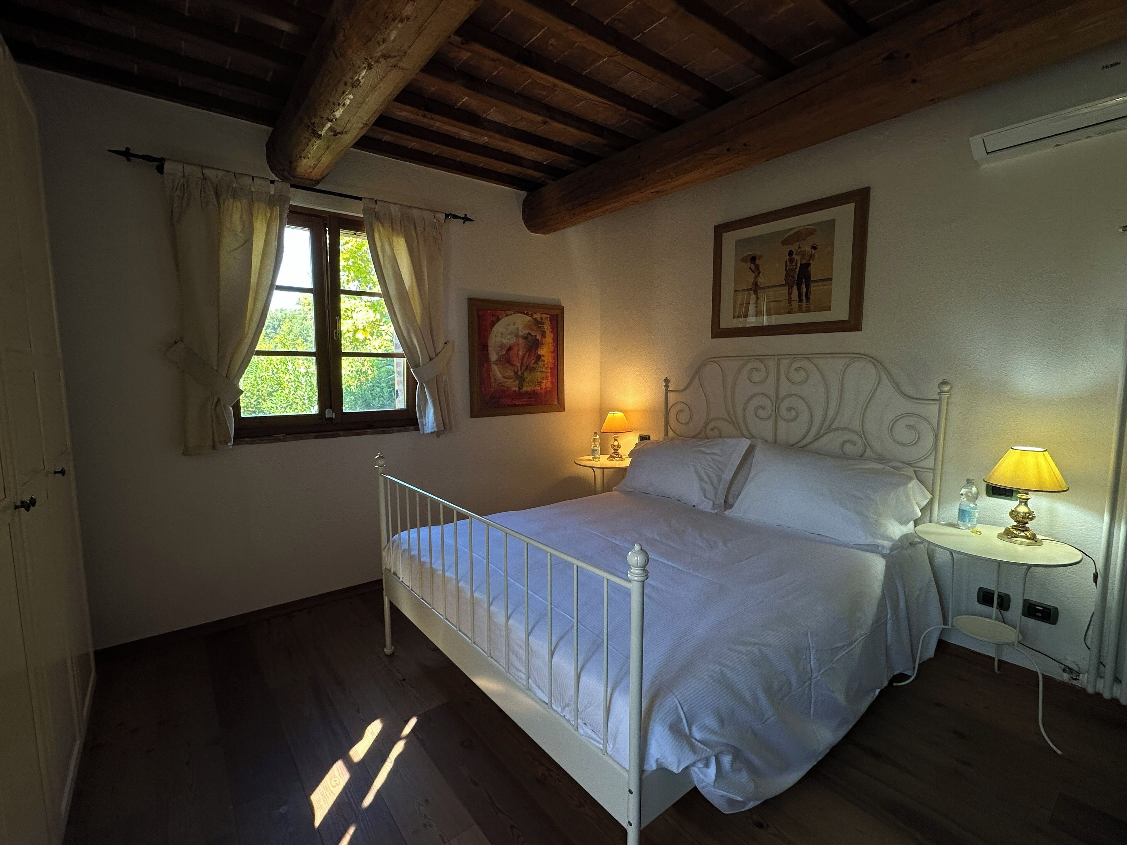 A bedroom with a bed, a window with curtains, a bedside table with a lamp, and a painting on the wall.