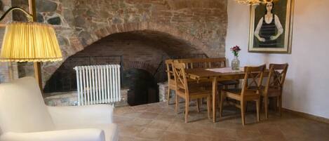 A stone fireplace, wooden beams, a white armchair, and a dining table with chairs.