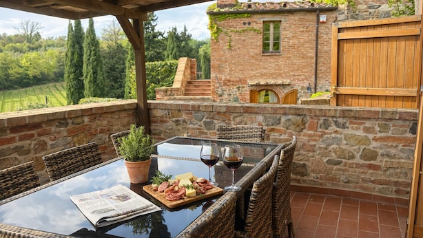 A covered outdoor dining area with wicker chairs, a glass table, and a view of a stone building with a wooden gate. - Agriturismo Borgo Sette Tigli (Montepulciano)
