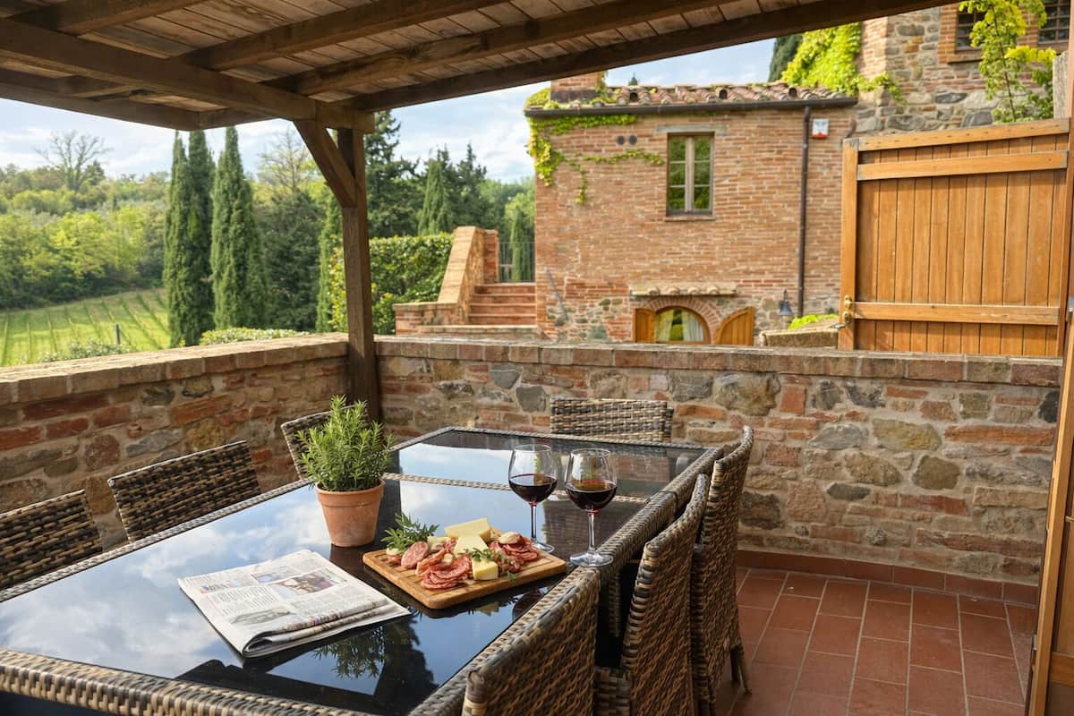 A covered outdoor dining area with wicker chairs, a glass table, and a view of a stone building with a wooden gate.