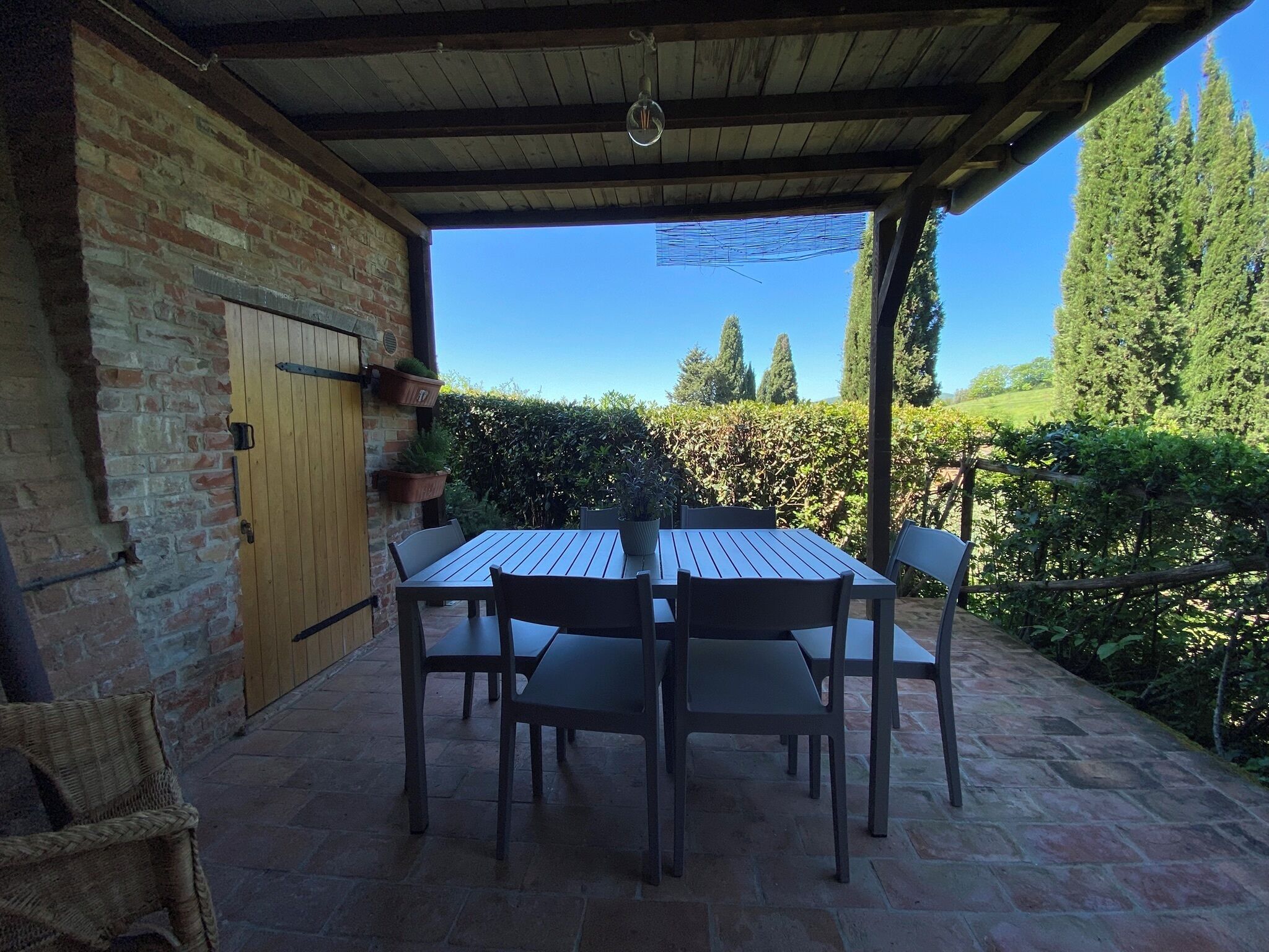 Um alpendre coberto com mesa e cadeiras de madeira, parede de tijolos e vista para um sebedo e árvores.