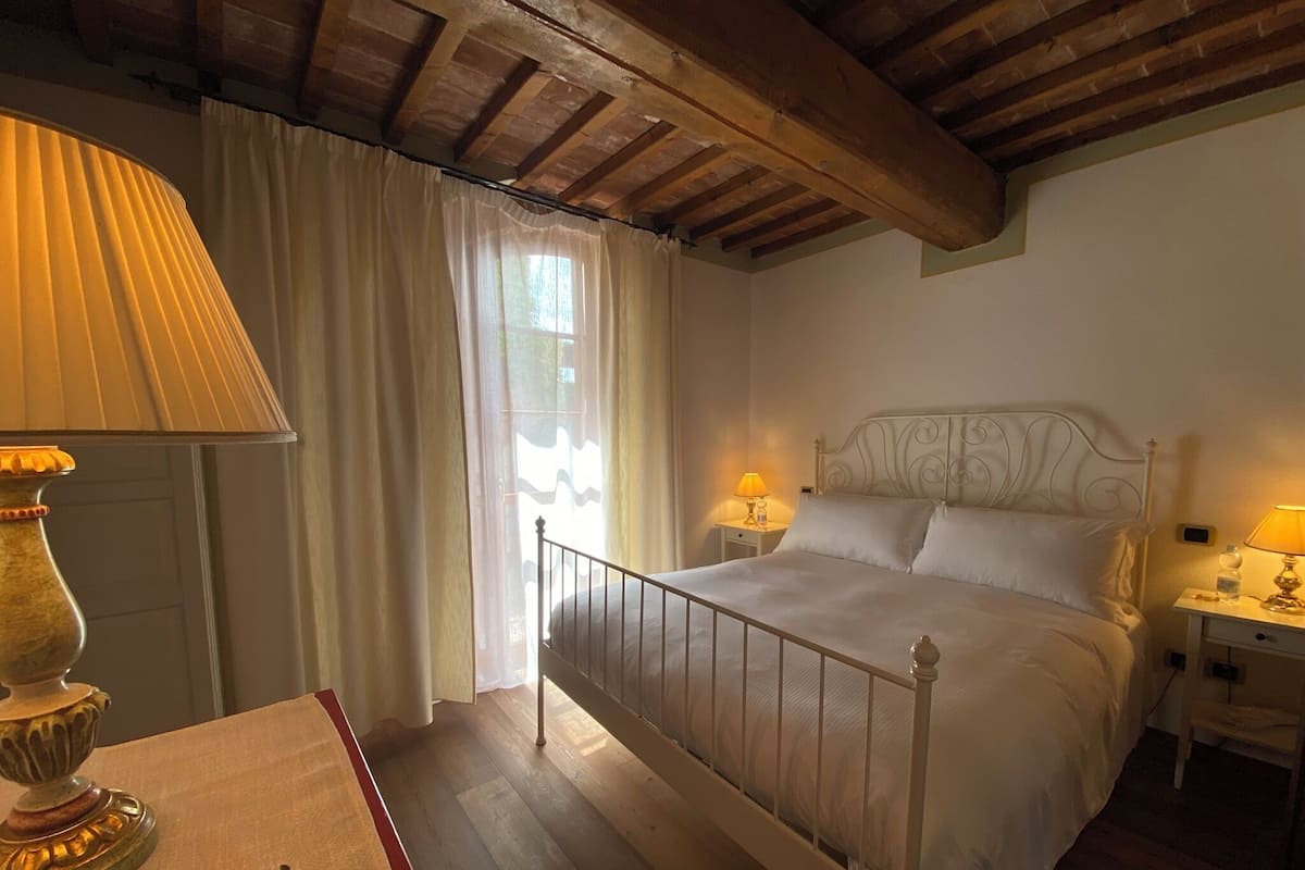 A bedroom with a wooden ceiling, a bed with white bedding, two bedside lamps, and a window with sheer curtains.