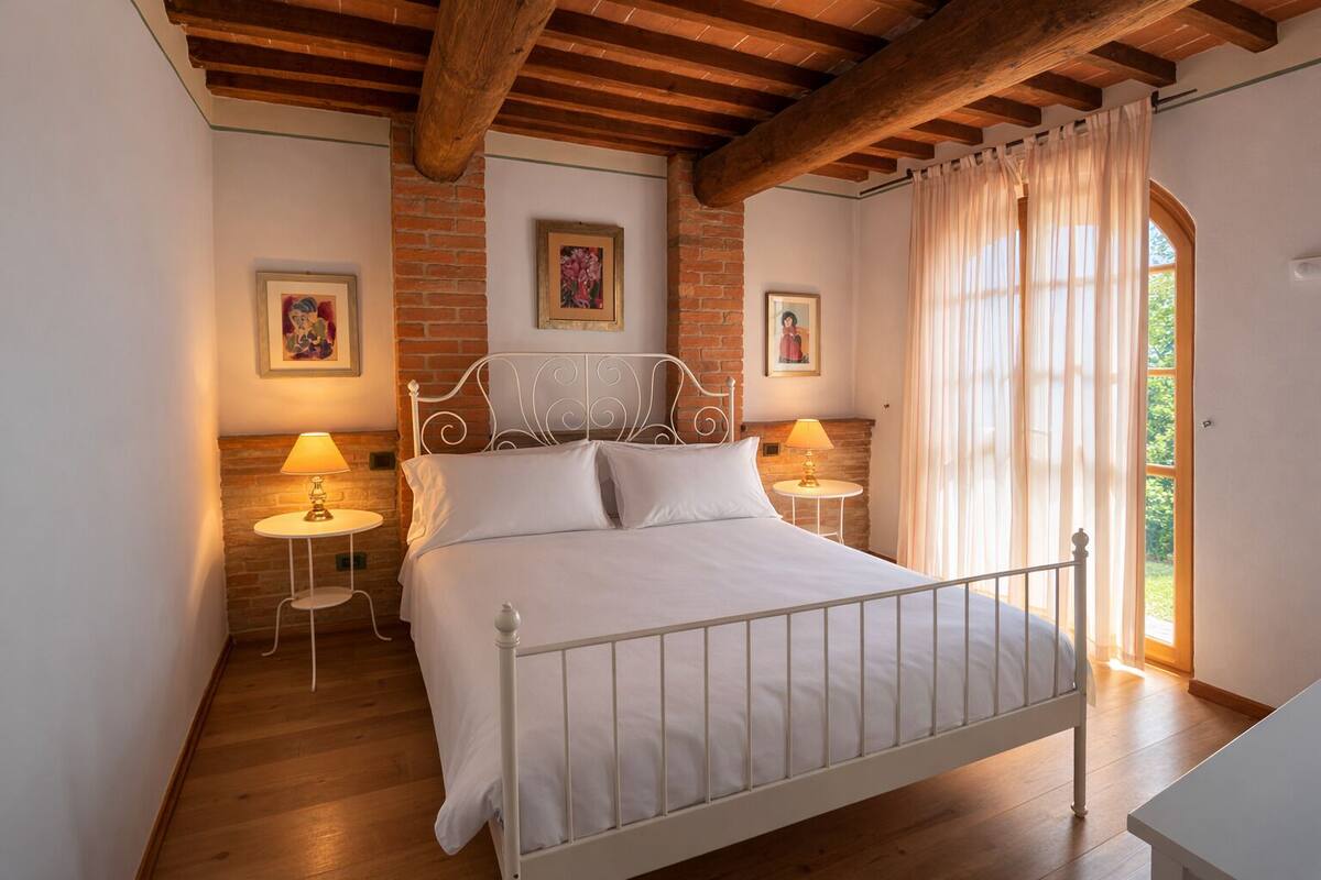 A bedroom with a white metal bed, two bedside tables with lamps, a brick wall, and a window with curtains.