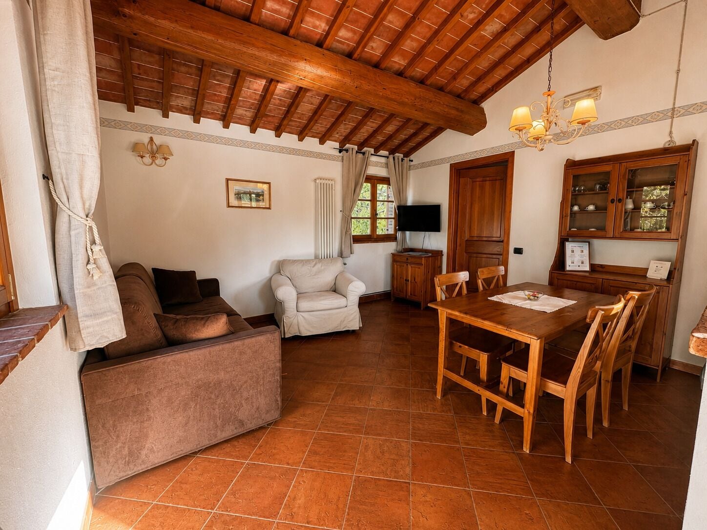 Uma sala de estar acolhedora com sofá, poltrona, mesa de jantar com cadeiras, teto de madeira e televisão sobre um móvel.