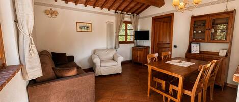 Uma sala de estar acolhedora com sofá, poltrona, mesa de jantar com cadeiras, teto de madeira e televisão sobre um móvel.