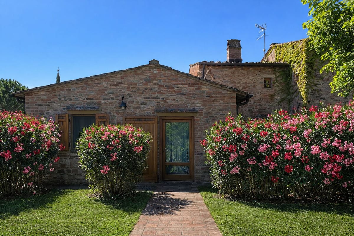 A stone building with a brick pathway leading to a wooden door, surrounded by lush pink flowering bushes.