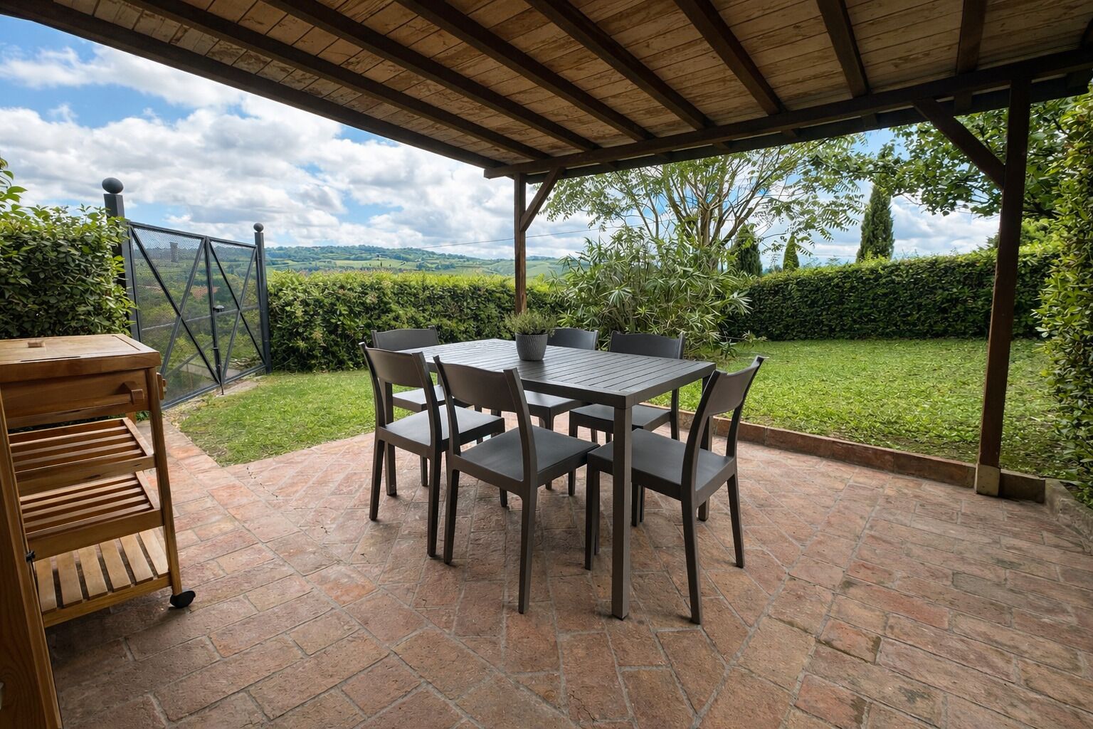 An outdoor patio with a table and chairs, a wooden cart, and a view of a green landscape.