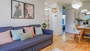 A modern living room with a grey sofa, colorful cushions, and two framed artworks. A wooden dining table with chairs is visible in the background. - Samantha Suite | City 2BR Bungalow Free Parking Garden & WIFI (Cardiff)
