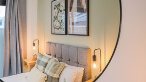 A bedroom with a grey upholstered headboard, a bed with white and green striped bedding, two framed artworks above the bed, and a nightstand with a lamp. - Samantha Suite | City 2BR Bungalow Free Parking Garden & WIFI (Cardiff)