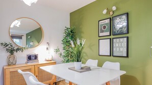A dining area with a white table, four white chairs, a wooden cabinet with a radio, a potted plant, and framed artwork on the wall. - Samantha Suite | City 2BR Bungalow Free Parking Garden & WIFI (Cardiff)