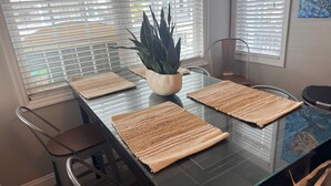 Uma sala de jantar moderna com mesa de vidro, jogos americanos trançados e um vaso com planta.