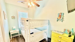 Quarto com beliche, ventoinha no teto, janela com persianas, mesinha com abajur, cômoda amarela e decorações nas paredes.