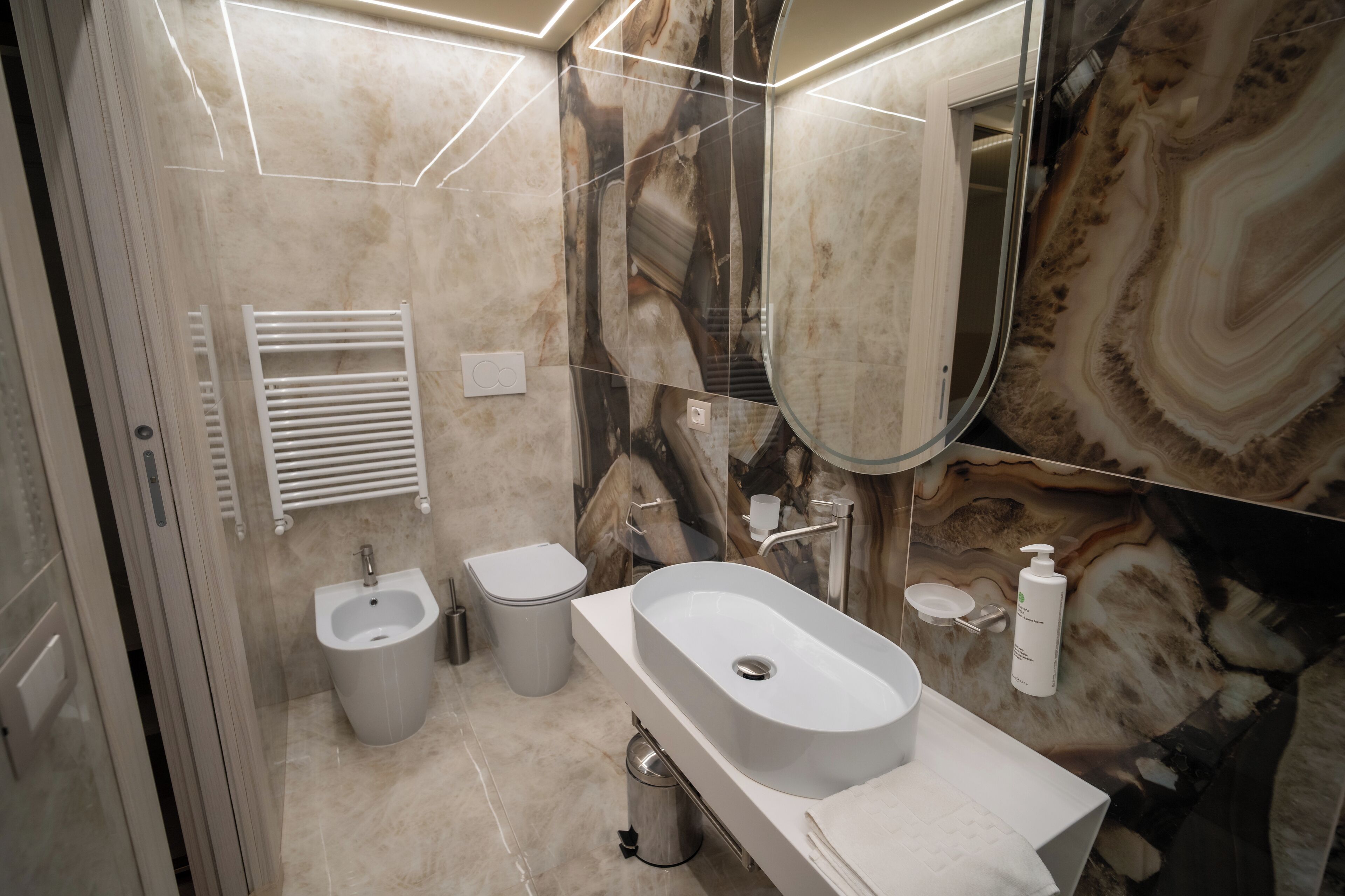 A modern bathroom with a marble backsplash, a large mirror, a sink, a bidet, and a toilet.