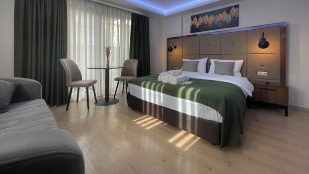 Una habitación de hotel moderna con una cama bien hecha, una pequeña mesa redonda con sillas y un ventanal con cortinas traslúcidas.