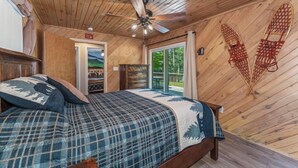 A wooden cabin bedroom with a bed, a ceiling fan, and a window with a view of trees. - Stepaway Vacation Rentals Loon's Hideaway on Pickerel Lake (St.Germain)