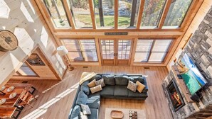 A spacious living room with a large sectional sofa, a fireplace, and large windows offering a view of a forested area.