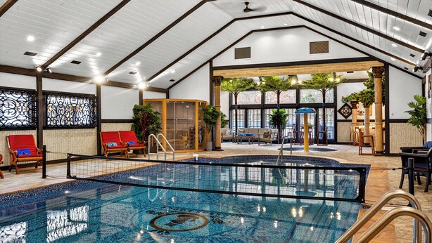 Una piscina interior con suelo de baldosas, rodeada de sillones de madera y una zona de estar con mesas y sillas.