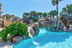 A pool area with a rock formation and a slide, surrounded by palm trees and buildings.