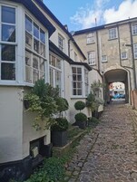 A narrow cobblestone street flanked by buildings with white-framed windows and a brick archway.